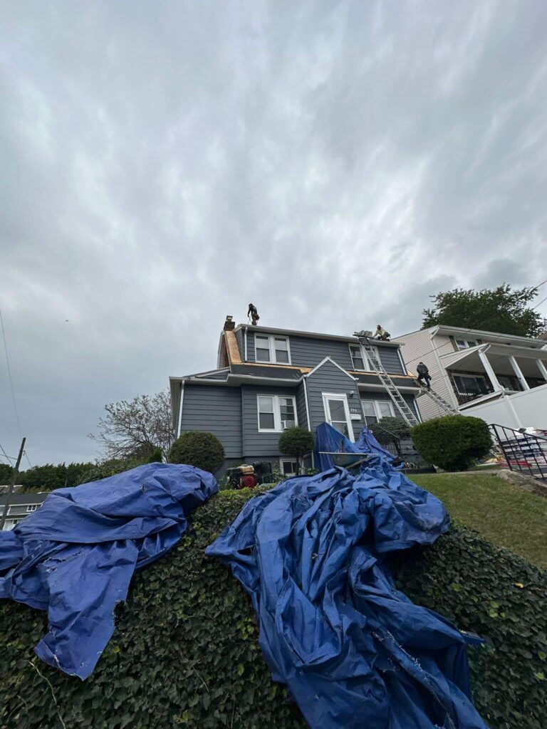 Roofers on a gray house with blue tarps on the ground, under a cloudy sky.