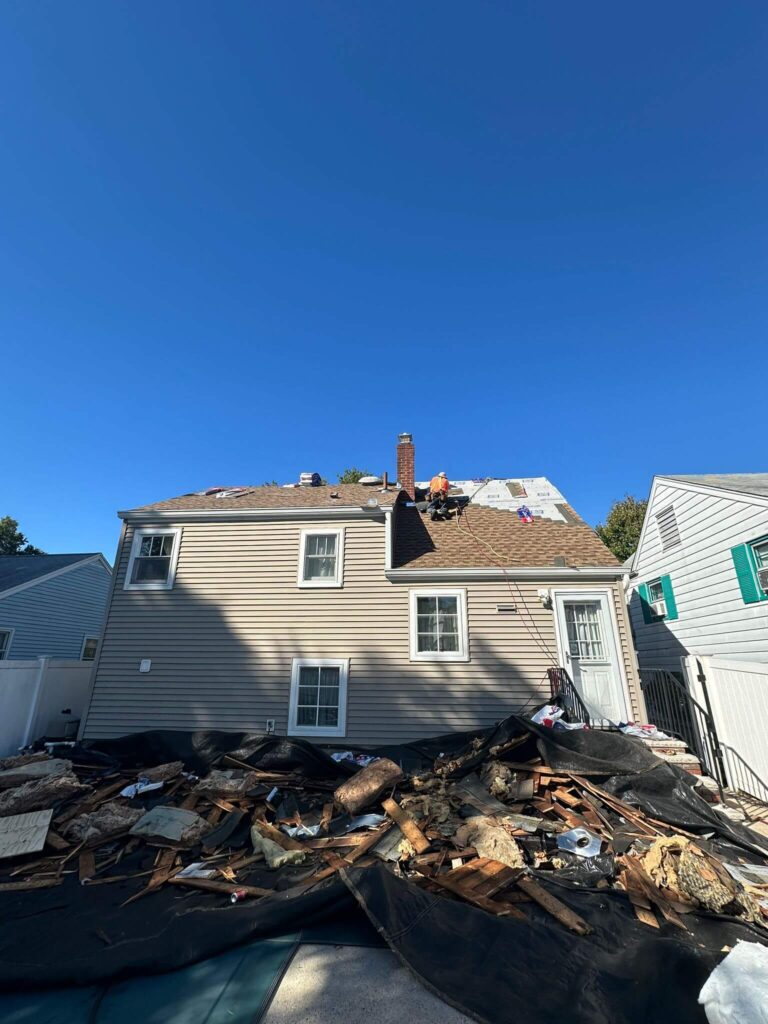 A roofer works on installing a new roof on a tan house with a pile of old roofing debris on a tarp below.