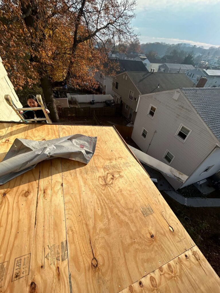 A roofer on a ladder next to a house with new plywood roof decking, with other houses and trees in the background.